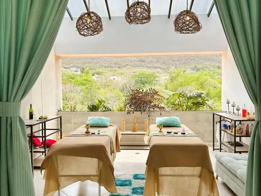 Sala del spa con dos camillas, cortinas verdes y el bosque al fondo