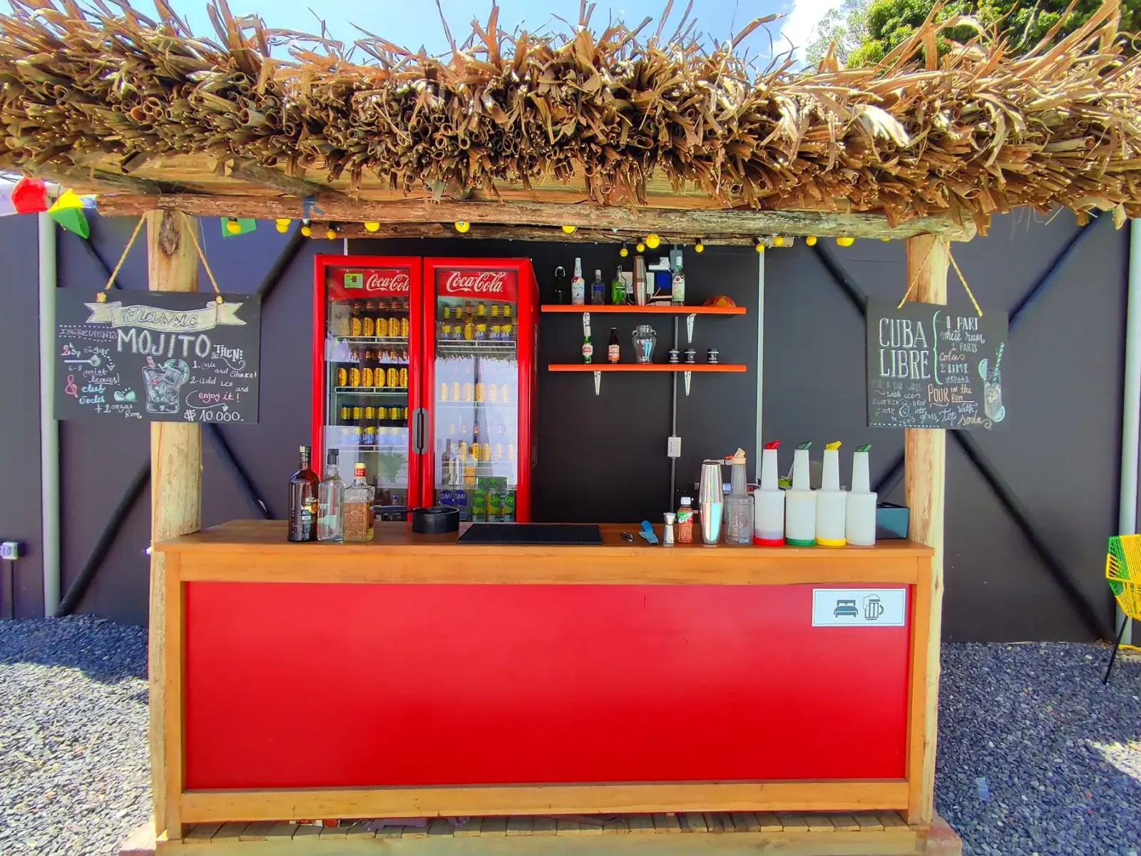 Barra roja con nevera, botellas y tableros de cócteles bajo un techo de palma en Origen