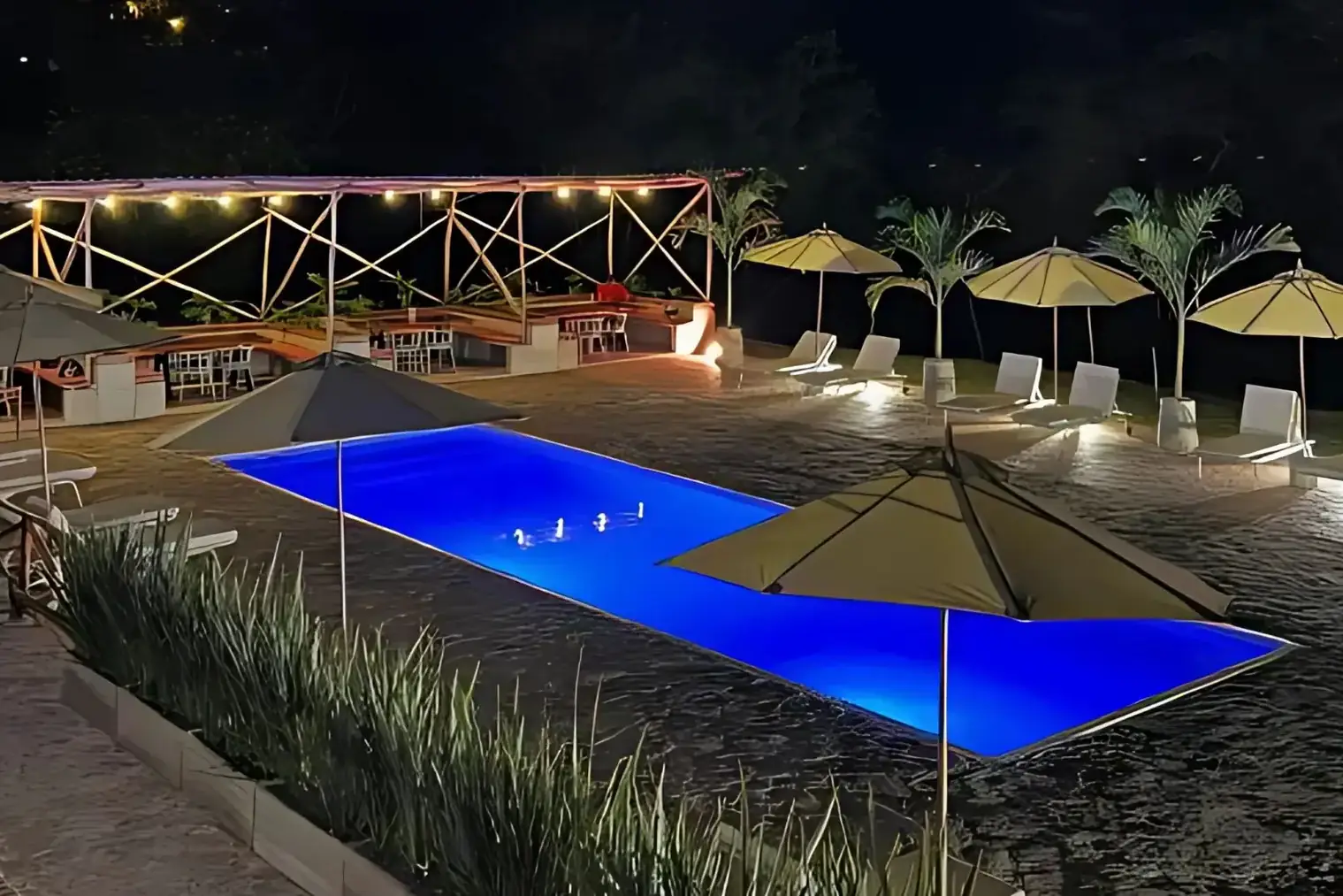 La piscina de Del Río iluminada de azul por la noche, con sombrillas cerradas alrededor